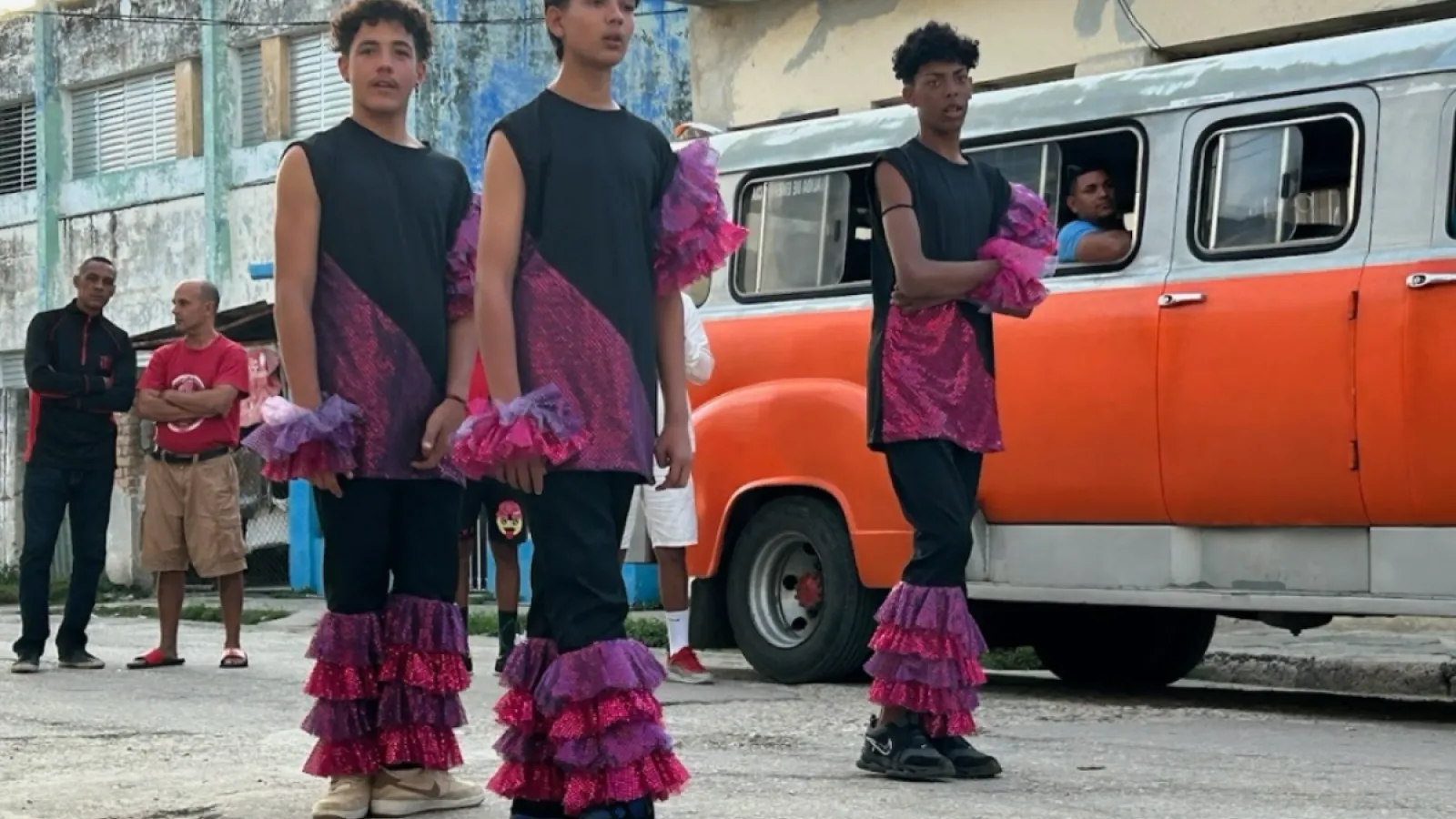 three men dancing wearing black clothing with pink ruffles in front of a red and white van