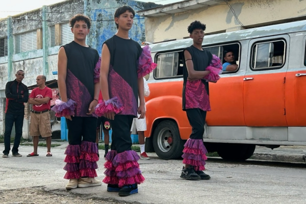 three men dancing wearing black clothing with pink ruffles in front of a red and white van